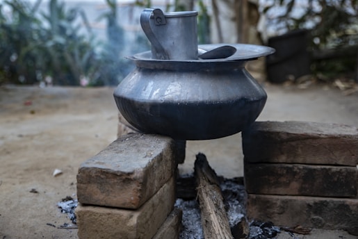 A metal pot sits atop a simple outdoor cooking setup, constructed using bricks and fueled by burning wood. Ashes and charred wood lie underneath the pot, while smoke rises slightly from the makeshift stove. The background is blurred and features greenery, suggesting a rural setting.