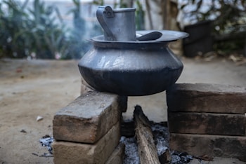 A metal pot sits atop a simple outdoor cooking setup, constructed using bricks and fueled by burning wood. Ashes and charred wood lie underneath the pot, while smoke rises slightly from the makeshift stove. The background is blurred and features greenery, suggesting a rural setting.