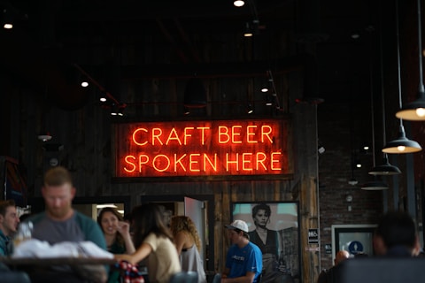 A dimly lit pub or bar setting featuring a neon sign that reads 'Craft Beer Spoken Here' in bright orange-red. A group of people are socializing and sitting at a wooden table in the foreground. The interior has a rustic ambiance with wooden panels and several hanging pendant lights. There is a black and white portrait on the wall adding to the decor.