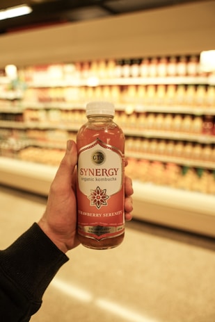 A hand is holding a bottle of GT's Synergy organic kombucha, labeled 'Strawberry Serenity,' in a grocery store aisle. Shelves filled with various products are blurred in the background, emphasizing the kombucha bottle.