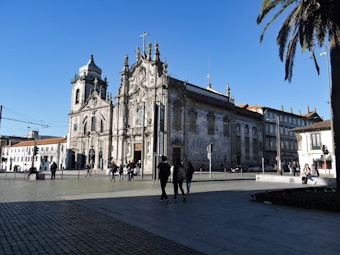 A historical, ornate church with intricate architectural details, tiled facades, and crosses on top. Several people are walking nearby, with a clear blue sky and palm trees adding to the urban scene.