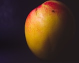 A close-up image of a ripe mango with a smooth yellow and red skin, highlighted against a dark background, enhancing its vivid colors and textures.