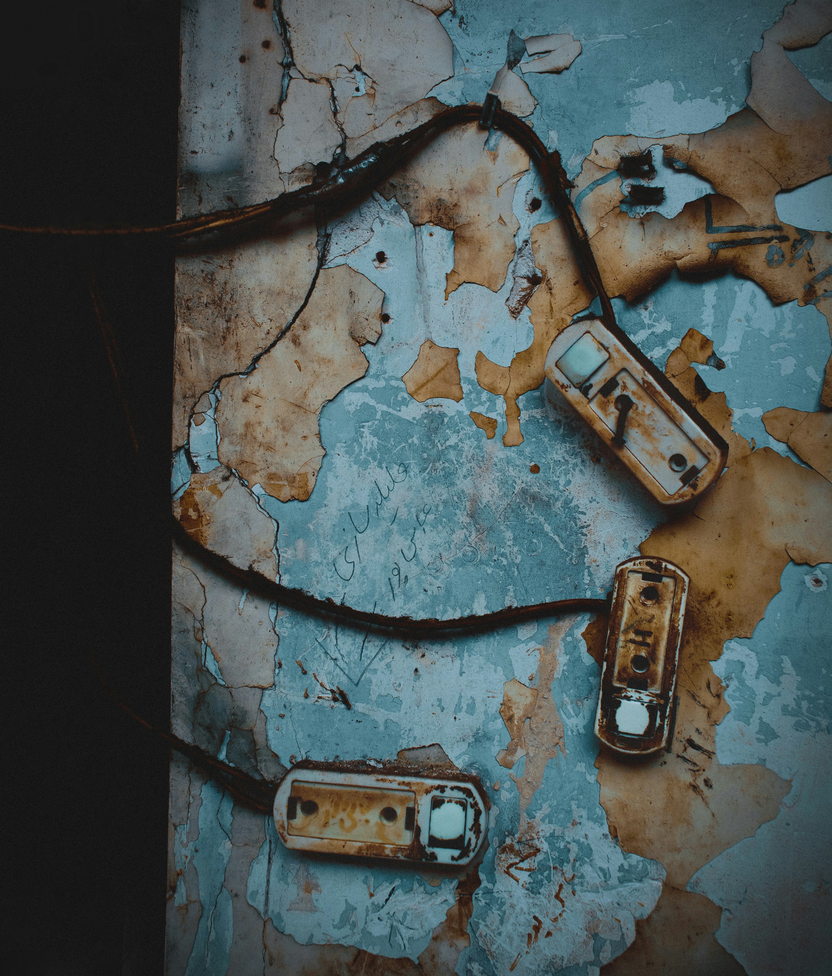 An old and peeling wall with a distressed appearance, featuring exposed wiring and three rusty light switches. The paint is worn and flaking off, revealing layers of history and neglect.