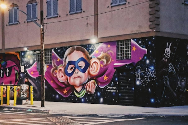 A vibrant mural featuring a child wearing a superhero mask with large, exaggerated ears, surrounded by a purple cape with yellow stars. The backdrop includes a cosmic space theme with stars and a dark sky. The art is on the side of a building with an urban street view and other graffiti art nearby.