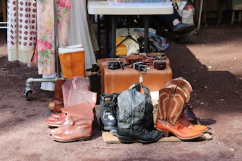 A collection of cowboy boots, leather shoes, and a variety of belts are displayed on the ground. In the background, clothing items with floral patterns and a table with more accessories are visible. Sunlight casts shadows on the ground, suggesting an outdoor market setting.