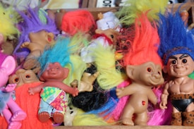 A collection of troll dolls with colorful hair is scattered, displaying a variety of vibrant colors like blue, purple, red, and yellow. Each doll has a distinctive look; some have clothes, while others are unclothed. The dolls vary in skin tones and expressions, adding to the visual diversity.