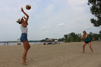 Two women are playing beach volleyball on a sandy beach with a lake in the background. One woman is jumping to hit the volleyball while the other stands ready to receive. The beach is populated with people enjoying the water and the outdoors. Trees line the background under a partly cloudy sky.