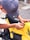 A person wearing a gray cap and a silver bracelet is applying a sticker to a bright yellow motorcycle. The motorcycle has a Honda logo and other branding elements on its body.