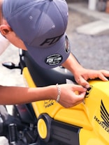 A person wearing a gray cap and a silver bracelet is applying a sticker to a bright yellow motorcycle. The motorcycle has a Honda logo and other branding elements on its body.