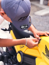 A person wearing a gray cap and a silver bracelet is applying a sticker to a bright yellow motorcycle. The motorcycle has a Honda logo and other branding elements on its body.