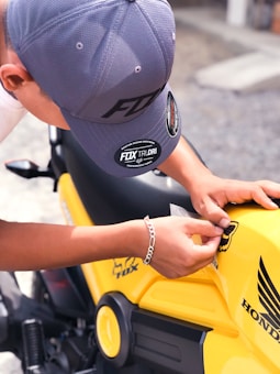 A person wearing a gray cap and a silver bracelet is applying a sticker to a bright yellow motorcycle. The motorcycle has a Honda logo and other branding elements on its body.