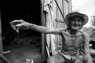 An elderly man with a weathered face is sitting in front of a wooden shed, reaching out with one arm. He wears a large brimmed hat and appears shirtless, showcasing a lean and muscular build. The open door of the shed reveals a shadowy interior with what appears to be some old fabric or bedding inside. Lush greenery can be seen in the background, suggesting a rural setting.