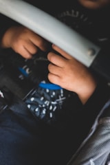A close-up view of a child's hands resting on a car seat, with a soft focus on the hands and a harness belt visible. The child's clothing features a patterned fabric.
