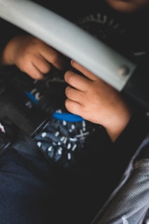 A close-up view of a child's hands resting on a car seat, with a soft focus on the hands and a harness belt visible. The child's clothing features a patterned fabric.