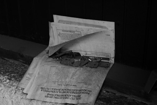 A stack of newspapers or papers is placed on a rough surface with several metal hinges and small parts on top. The scene is in black and white, creating a vintage or nostalgic feel. The background surface is dark, possibly a wooden bench or shelf, while the papers are older, possibly instruction manuals in various languages.