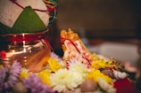 A small Ganesha idol is surrounded by vibrant yellow and white flowers, placed beside a traditional Kalash with a coconut on top and adorned with a green leaf. The setting suggests a ceremonial or religious context.