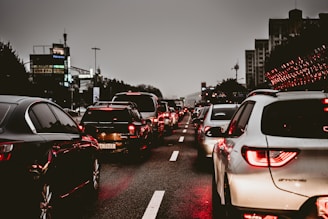 Heavy traffic on a multi-lane road during an overcast evening. Numerous cars are lined up, displaying bright red taillights as they are either stopped or moving slowly. The surrounding environment includes city buildings and streetlights, contributing to an urban setting.