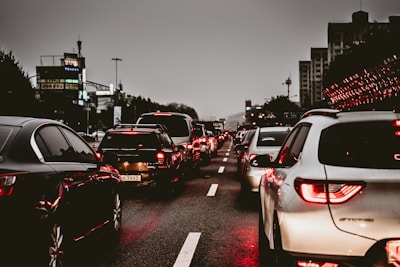 Heavy traffic on a multi-lane road during an overcast evening. Numerous cars are lined up, displaying bright red taillights as they are either stopped or moving slowly. The surrounding environment includes city buildings and streetlights, contributing to an urban setting.