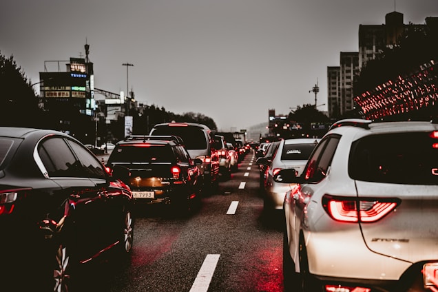 Heavy traffic on a multi-lane road during an overcast evening. Numerous cars are lined up, displaying bright red taillights as they are either stopped or moving slowly. The surrounding environment includes city buildings and streetlights, contributing to an urban setting.