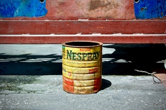 A vintage Nestlé Nido can, used as a container, is placed on a concrete surface. The can is rusted and weathered, with faded colors and visible brand markings. It stands against a backdrop of a red wooden surface with peeling blue paint.