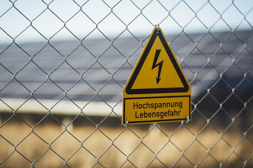 A triangular yellow warning sign with a black lightning bolt icon and the text 'Hochspannung Lebensgefahr' in black is attached to a wire fence. The background shows blurred solar panels and greenery, suggesting a location with potential electrical hazards.