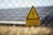 A triangular yellow warning sign with a black lightning bolt icon and the text 'Hochspannung Lebensgefahr' in black is attached to a wire fence. The background shows blurred solar panels and greenery, suggesting a location with potential electrical hazards.