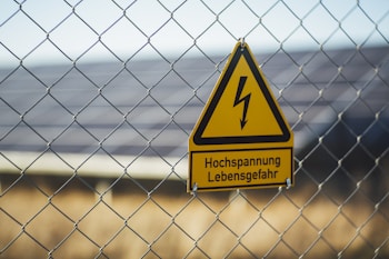 A triangular yellow warning sign with a black lightning bolt icon and the text 'Hochspannung Lebensgefahr' in black is attached to a wire fence. The background shows blurred solar panels and greenery, suggesting a location with potential electrical hazards.