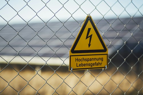 A triangular yellow warning sign with a black lightning bolt icon and the text 'Hochspannung Lebensgefahr' in black is attached to a wire fence. The background shows blurred solar panels and greenery, suggesting a location with potential electrical hazards.
