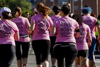 A group of women wearing matching pink shirts with text on the back, participating in a run or charity event. They are seen from behind, running together on a sunny day with trees and buildings in the background.
