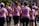 A group of women wearing matching pink shirts with text on the back, participating in a run or charity event. They are seen from behind, running together on a sunny day with trees and buildings in the background.