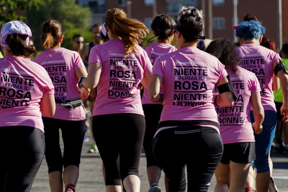 A group of women wearing matching pink shirts with text on the back, participating in a run or charity event. They are seen from behind, running together on a sunny day with trees and buildings in the background.