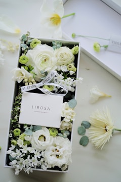 A beautifully arranged box of white and light green flowers surrounding a product box labeled 'LIROSA Natural Gel Cream'. Delicate blossoms including roses and various small blooms are interspersed with green foliage. The arrangement is accented with ribbons labeled 'VIOLET'. Scattered petals and flowers are placed outside the box on a light-colored table.