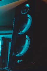 A close-up view of a black speaker featuring two large circular drivers. The speaker is standing upright in a dimly lit environment with a bluish tint, highlighting its textured surface and the brand logo near the bottom. The top driver is smaller, with distinct concentric patterns visible in the recessed areas of the drivers.
