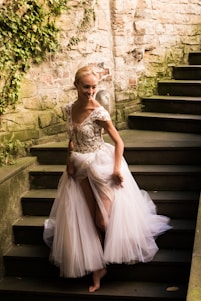 A person wearing an elegant, flowing gown with intricate floral embroidery on the bodice walks down a stone staircase. The stone wall is partially covered in greenery, adding a natural element to the setting.