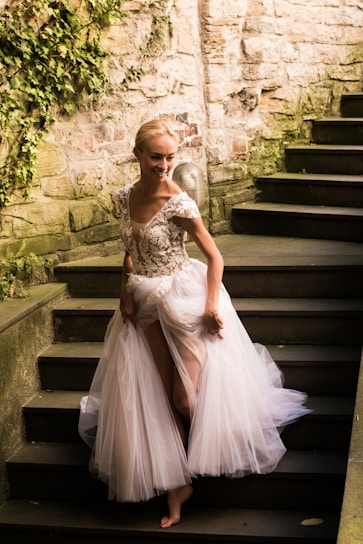 A person wearing an elegant, flowing gown with intricate floral embroidery on the bodice walks down a stone staircase. The stone wall is partially covered in greenery, adding a natural element to the setting.