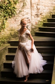 A person wearing an elegant, flowing gown with intricate floral embroidery on the bodice walks down a stone staircase. The stone wall is partially covered in greenery, adding a natural element to the setting.