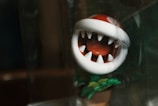 A small, toy-like figure of a stylized plant with a large mouth and sharp, white teeth. Its head is predominantly red with white spots, reminiscent of a piranha plant. The figure appears to be inside a transparent container, and green leaves are visible at the base.