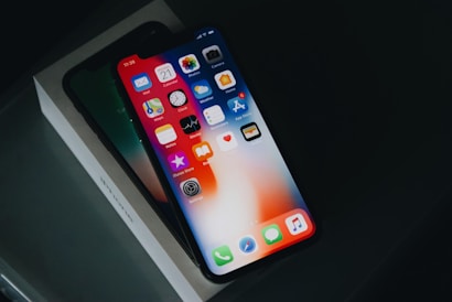 A modern smartphone displaying its home screen with various app icons is placed at an angle on top of its box, emphasizing its sleek design against a dark background.