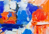 Thick, textured paint in vibrant shades of blue, orange, and white is spread across a canvas. The brushstrokes are heavy and layered, creating a dynamic, abstract composition.