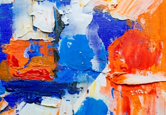 Thick, textured paint in vibrant shades of blue, orange, and white is spread across a canvas. The brushstrokes are heavy and layered, creating a dynamic, abstract composition.