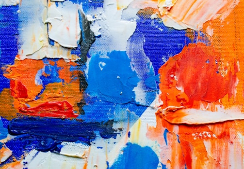 Thick, textured paint in vibrant shades of blue, orange, and white is spread across a canvas. The brushstrokes are heavy and layered, creating a dynamic, abstract composition.
