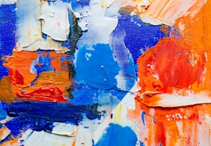 Thick, textured paint in vibrant shades of blue, orange, and white is spread across a canvas. The brushstrokes are heavy and layered, creating a dynamic, abstract composition.