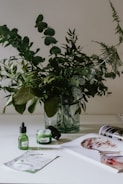 A glass vase holds an arrangement of lush green leafy branches on a bright, minimalistic surface. In the foreground, skincare products in green containers are positioned next to an open magazine displaying a colorful portrait. The overall setting conveys a fresh, natural aesthetic.