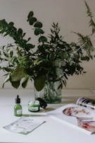 A glass vase holds an arrangement of lush green leafy branches on a bright, minimalistic surface. In the foreground, skincare products in green containers are positioned next to an open magazine displaying a colorful portrait. The overall setting conveys a fresh, natural aesthetic.