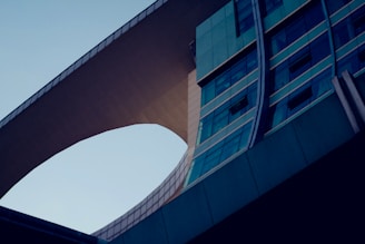 A modern architectural structure with smooth curves and a bridge-like feature connecting two sections of the building. The design incorporates extensive use of glass panels and steel, creating a reflective and sleek appearance.