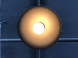 A circular ceiling light fixture emits a warm, glowing light in a darkened environment. The brushed metal surface reflects a gradient of light from bright white at the center to darker shades of bronze or gold towards the edges. Behind the light is a crossed pattern of pipes or bars, running vertically and horizontally across a textured, gray concrete wall.