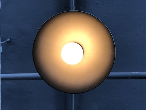 A circular ceiling light fixture emits a warm, glowing light in a darkened environment. The brushed metal surface reflects a gradient of light from bright white at the center to darker shades of bronze or gold towards the edges. Behind the light is a crossed pattern of pipes or bars, running vertically and horizontally across a textured, gray concrete wall.