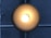 A circular ceiling light fixture emits a warm, glowing light in a darkened environment. The brushed metal surface reflects a gradient of light from bright white at the center to darker shades of bronze or gold towards the edges. Behind the light is a crossed pattern of pipes or bars, running vertically and horizontally across a textured, gray concrete wall.