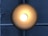 A circular ceiling light fixture emits a warm, glowing light in a darkened environment. The brushed metal surface reflects a gradient of light from bright white at the center to darker shades of bronze or gold towards the edges. Behind the light is a crossed pattern of pipes or bars, running vertically and horizontally across a textured, gray concrete wall.