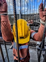 A construction worker wearing a yellow safety helmet and orange protective gloves is holding onto vertical metal rods. The person is dressed in a long-sleeved high-visibility shirt. The scene appears to be on a construction site with a cloudy sky and structural elements visible in the background.